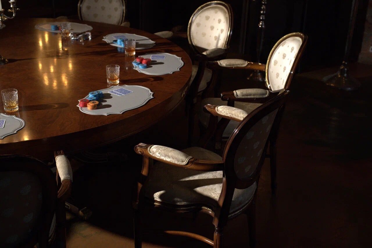 Арендовать Polygon(Полигон). Зал Апартаменты. Круглый деревянный стол с деревянными стульями с обивкой светлого цвета. Несколько стеклянных стаканов, настольные подставки с картами и фишками. Тёмное освещение, тёмный пол, минимум декора.