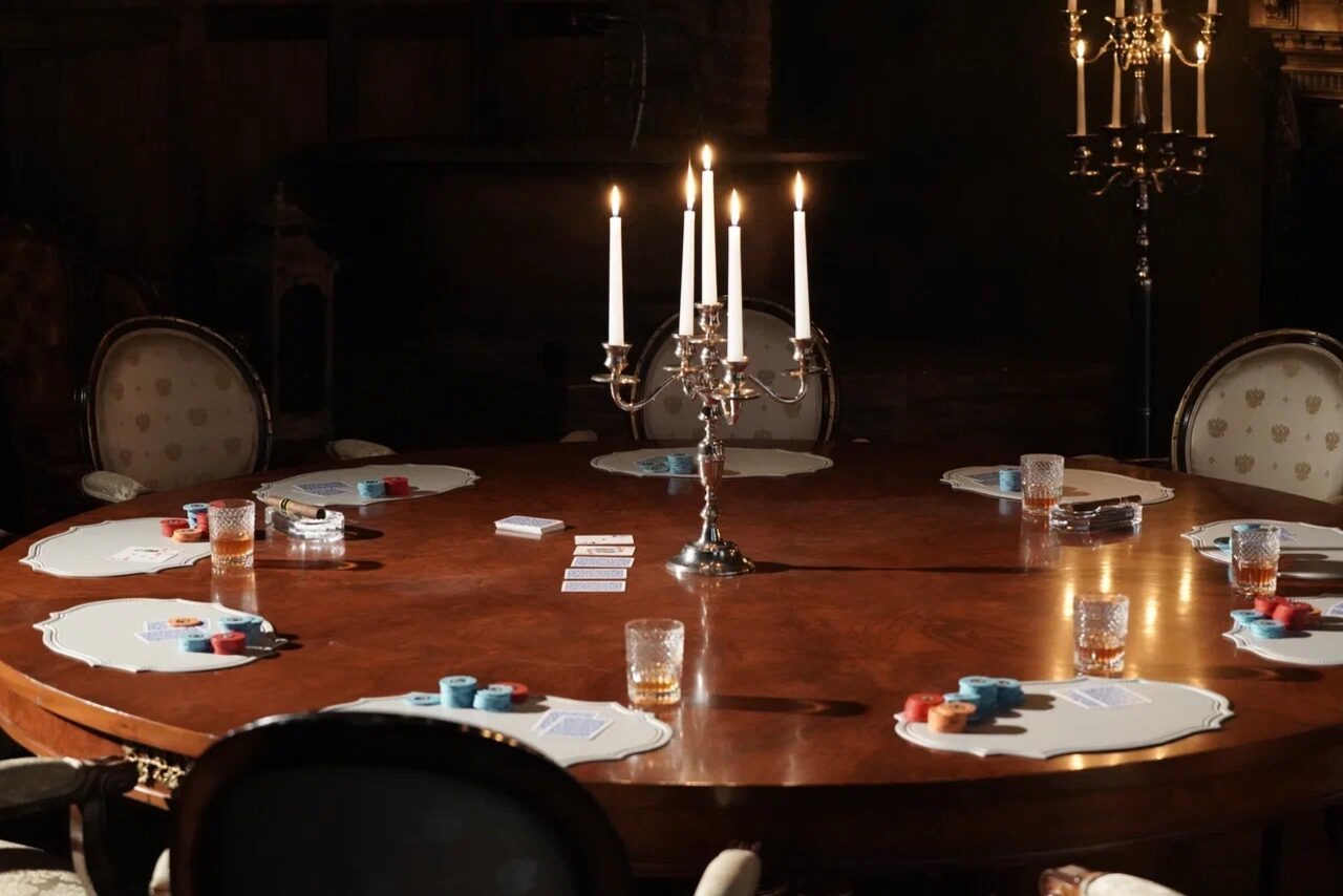 Арендовать Polygon(Полигон). Зал Апартаменты. Круглый деревянный стол с подсвечником по центру, окруженный стульями. На столе представлены подставки, игральные карты и фишки. Обстановка дополняется бокалами и свечами на подсвечниках, обеспечивающими мягкое освещение. Цвета темные, детали классические.