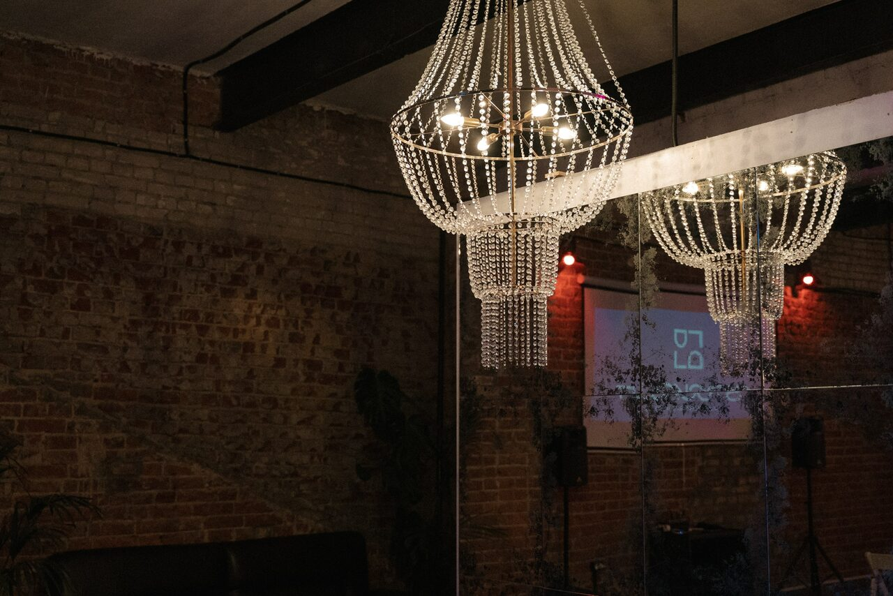 Арендовать Лофт с высокими потолками на востоке Москвы. Пространство с кирпичными стенами, зеркалом и черными балками под потолком. Потолочные люстры с кристаллами, пушистое растение рядом с темной мебелью. На стене проектор с изображением и колонка. Освещение теплое, доминирует темная цветовая гамма.