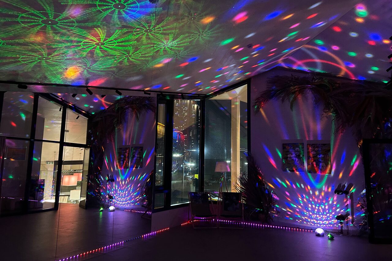 Арендовать Просторный зал с зеркалами для тренировок и мероприятий. Просторное помещение с высокими потолками и большими окнами; разноцветные световые проекции на стенах и потолке; стеклянные двери; несколько растений вдоль стен; видны картины; на полу тонкие светодиодные ленты.