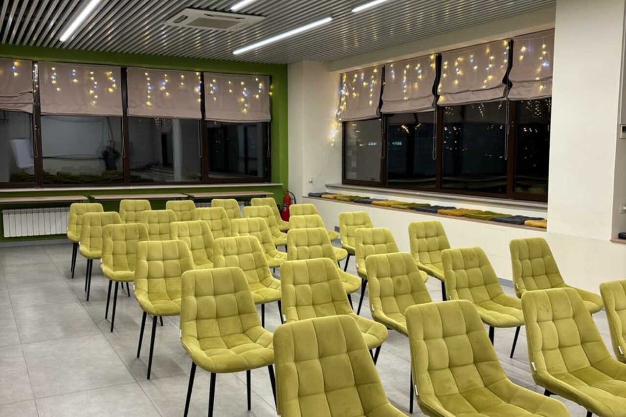 Арендовать Зал 6. Просторный и светлый конференц-зал до 50 человек в Академии Кантаты. Конференц-зал с рядами светло-зеленых стульев на черных ножках, светлые плиточные полы, большие окна с шторами, украшенные светящимися гирляндой, белые стены с зелеными акцентами, потолочные лампы, кондиционер в потолке.