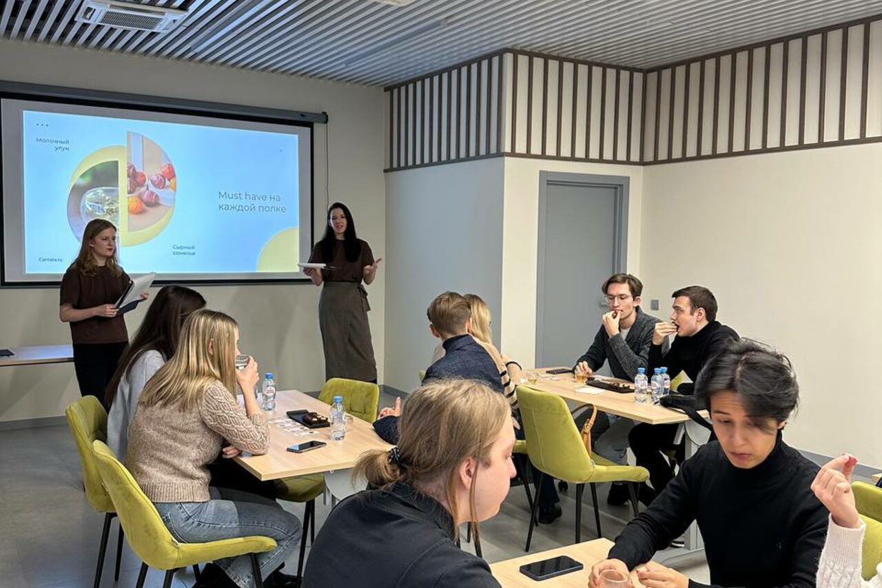 Арендовать Зал 6. Просторный и светлый конференц-зал до 50 человек в Академии Кантаты. Современное офисное пространство с людьми за столами. Стены светлые, потолок с решеткой. Мебель: небольшие деревянные столы, желтые стулья. Зона презентации с проектором и экраном. На экране текст и изображения. Все присутствующие заняты обсуждением.