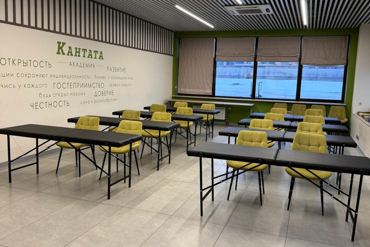 Арендовать Зал 6. Просторный и светлый конференц-зал до 50 человек в Академии Кантаты. Аудитория с рядами черных столов и желтыми стульями. На стене текст с надписью "КАНТАТА". Окно с видом на воду; жалюзи. Потолок с ламелями и встроенными светильниками. Светлая плитка на полу.