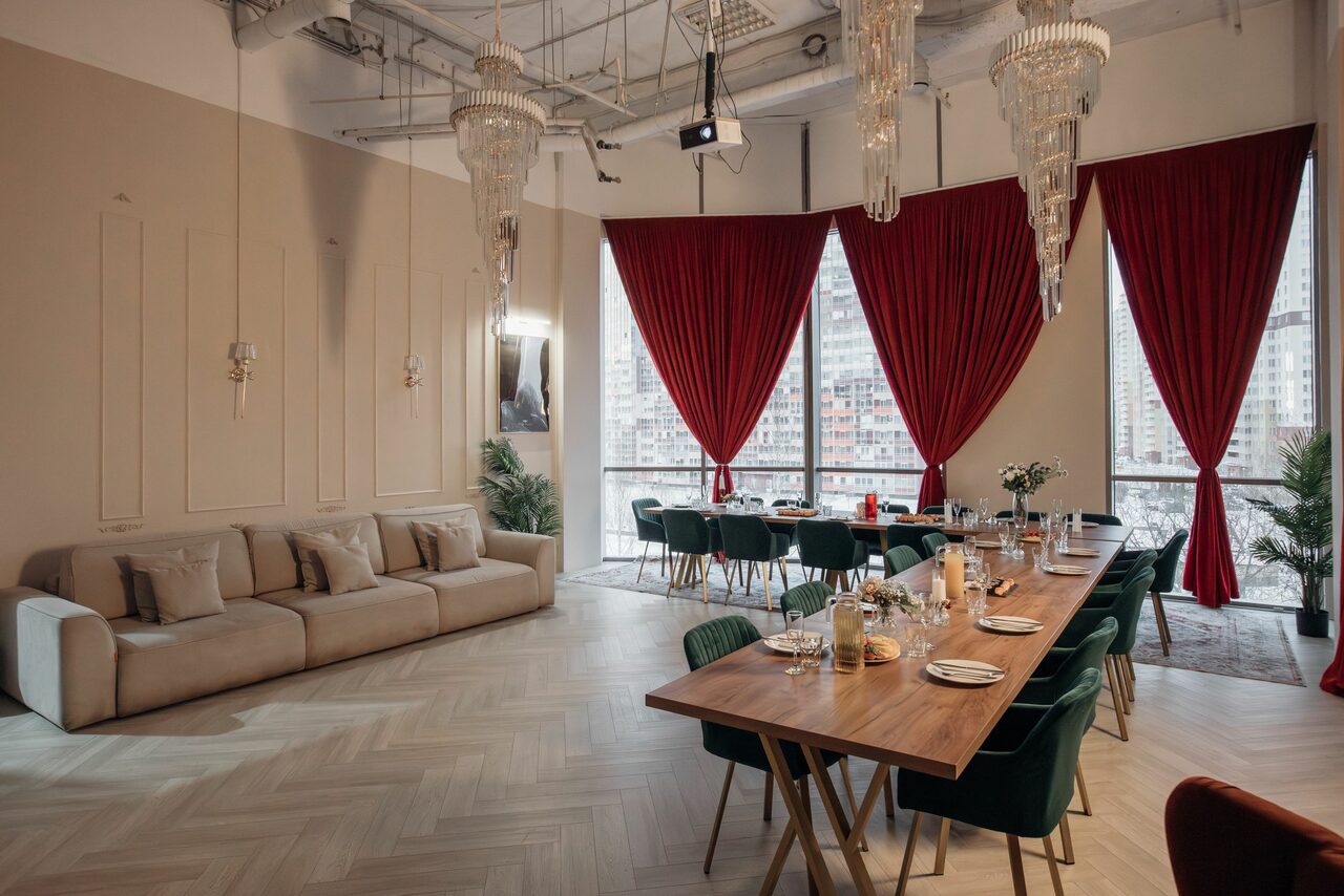 Арендовать Rubin Loft (Рубин Лофт). Зал Олд Мани. Просторное помещение с высокими потолками, большим деревянным столом и стульями, бежевым диваном. Красные шторы на больших окнах. Светлые паркетные полы, декоративные панели на стенах. Потолок с обнаженными коммуникациями и подвесными люстрами.