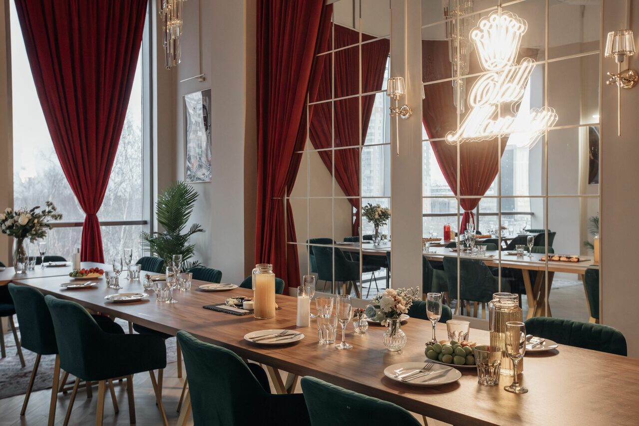 Арендовать Rubin Loft (Рубин Лофт). Зал Олд Мани. Большое помещение с длинным деревянным столом, окруженным мягкими стульями. На столе посуда и декор. Зеркальные стены с красными шторами и надпись в неоне. Естественное освещение из высоких окон. Декоративные растения.