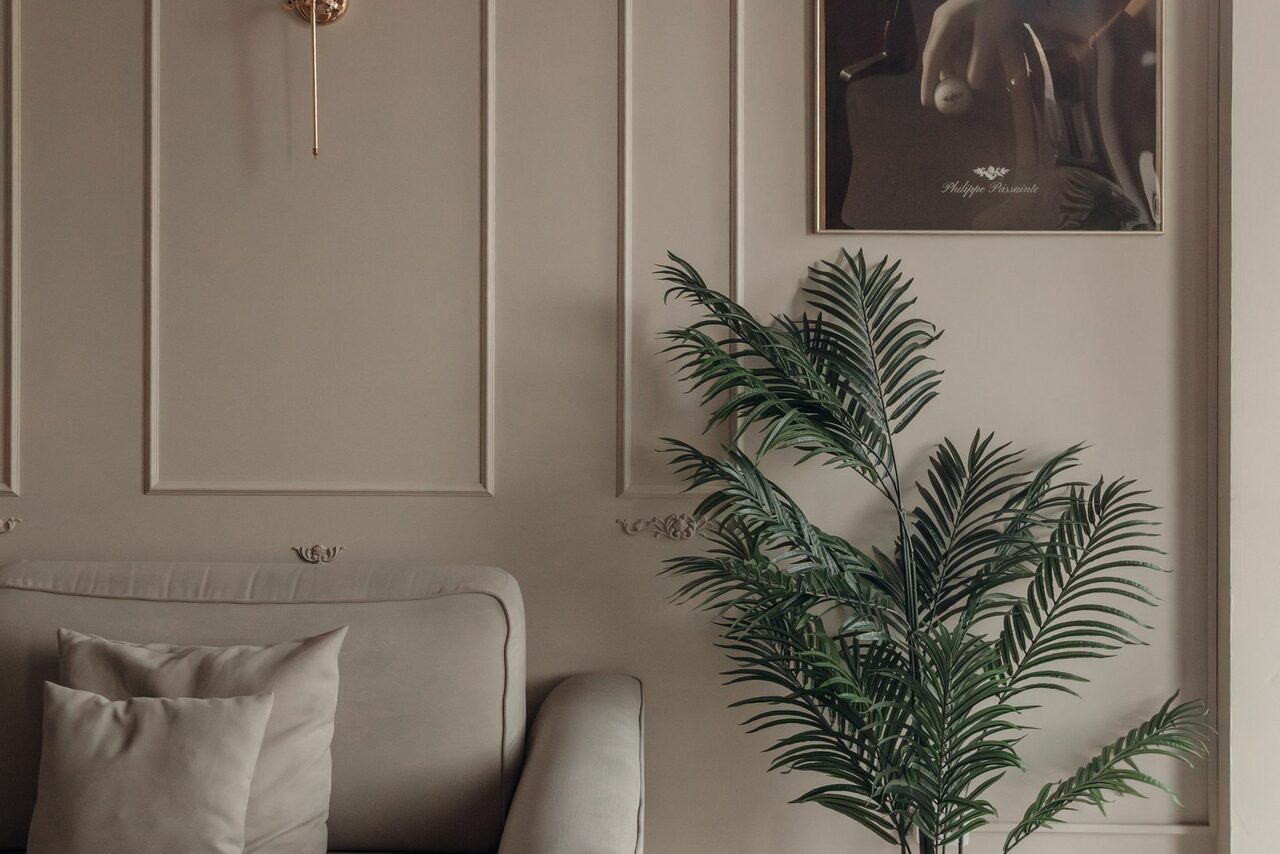 Арендовать Rubin Loft (Рубин Лофт). Зал Олд Мани. Гостиная с бежевыми стенами, мягким креслом в том же цвете и декоративными подушками. На стене золотистый бра и картина. Напольное растение с зелеными листьями. Стены с панелями, классический стиль. Теплое освещение.