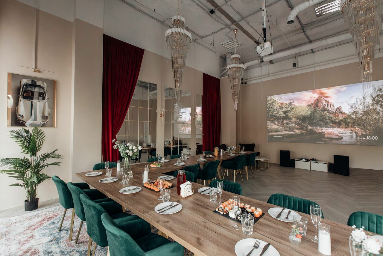 Арендовать Rubin Loft (Рубин Лофт). Зал Олд Мани. Просторное помещение с высоким потолком и открытыми коммуникациями. Длинный деревянный стол с зелеными стульями. Красные шторы, большие зеркала, проектор с изображением пейзажа, декоративные элементы, люстры, растения. Светлые стены, паркетный пол.