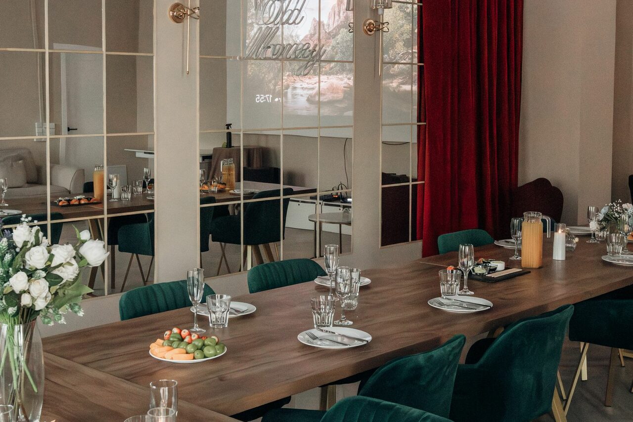 Арендовать Rubin Loft (Рубин Лофт). Зал Олд Мани. Пространство с длинным деревянным столом, окружённым стульями с зелёной обивкой. На столе стоят цветы, напитки и закуски. Задняя стена с зеркальными частями и проектором. Красные шторы. Бежевые стены. Посуду сервированы.