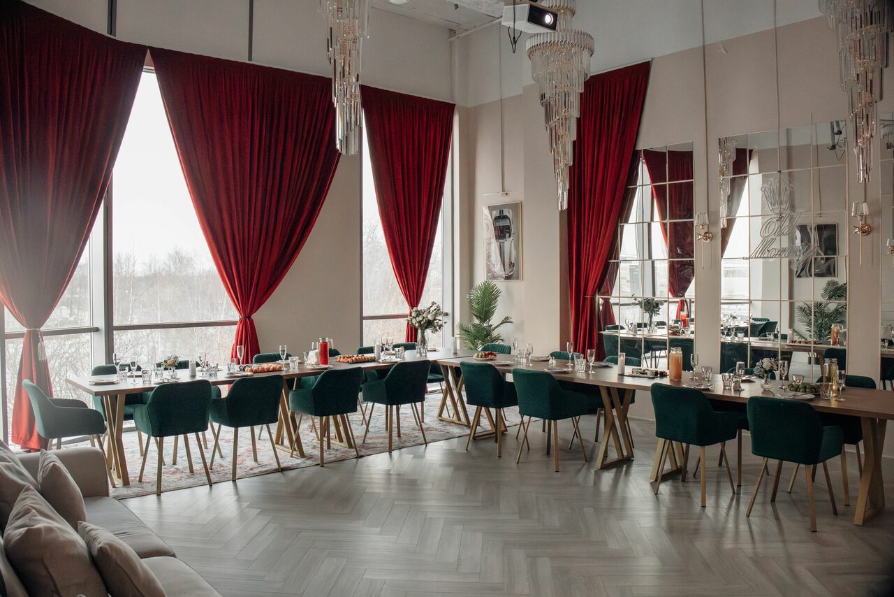 Арендовать Rubin Loft (Рубин Лофт). Зал Олд Мани. Просторное помещение с большими окнами, закрытыми красными шторами. Длинные столы с сервировкой, вокруг темно-зеленые стулья. На полу паркет светлого оттенка. Потолочные хрустальные люстры. Белые стены, на них большие зеркала.