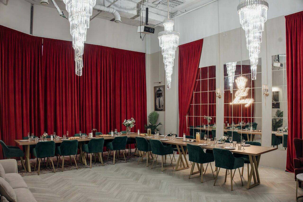 Арендовать Rubin Loft (Рубин Лофт). Зал Олд Мани. Просторное помещение с длинными деревянными столами и зелёными креслами. На стенах зеркальные панели, декорированные неоновыми надписями. Яркие красные занавески и белые люстры. Паркетный пол в светлых тонах, ваза с цветами на столе.