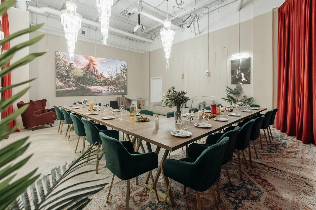Арендовать Rubin Loft (Рубин Лофт). Зал Олд Мани. Просторная комната с длинным деревянным столом и зелеными стульями на металлических ножках. На столе посуда и напитки. Светильники с подвесными кристаллами. Окружение дополнено красными шторами, картинами, ковром и живыми растениями. Светлые стены и высокий потолок.