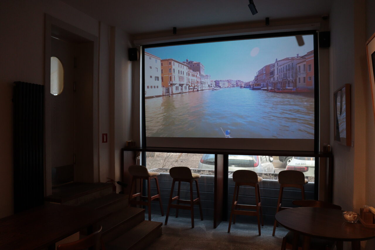 Арендовать Гастробар в бутик-отеле MOLO(Моло) 4. Небольшое помещение с низким потолком, оборудованное проектором и экраном, на котором изображена венецианская сцена. Рядом стоят четыре деревянных стула у окна. В помещении также видны круглые столы и лестница. Освещение мягкое. Пол ковровый.
