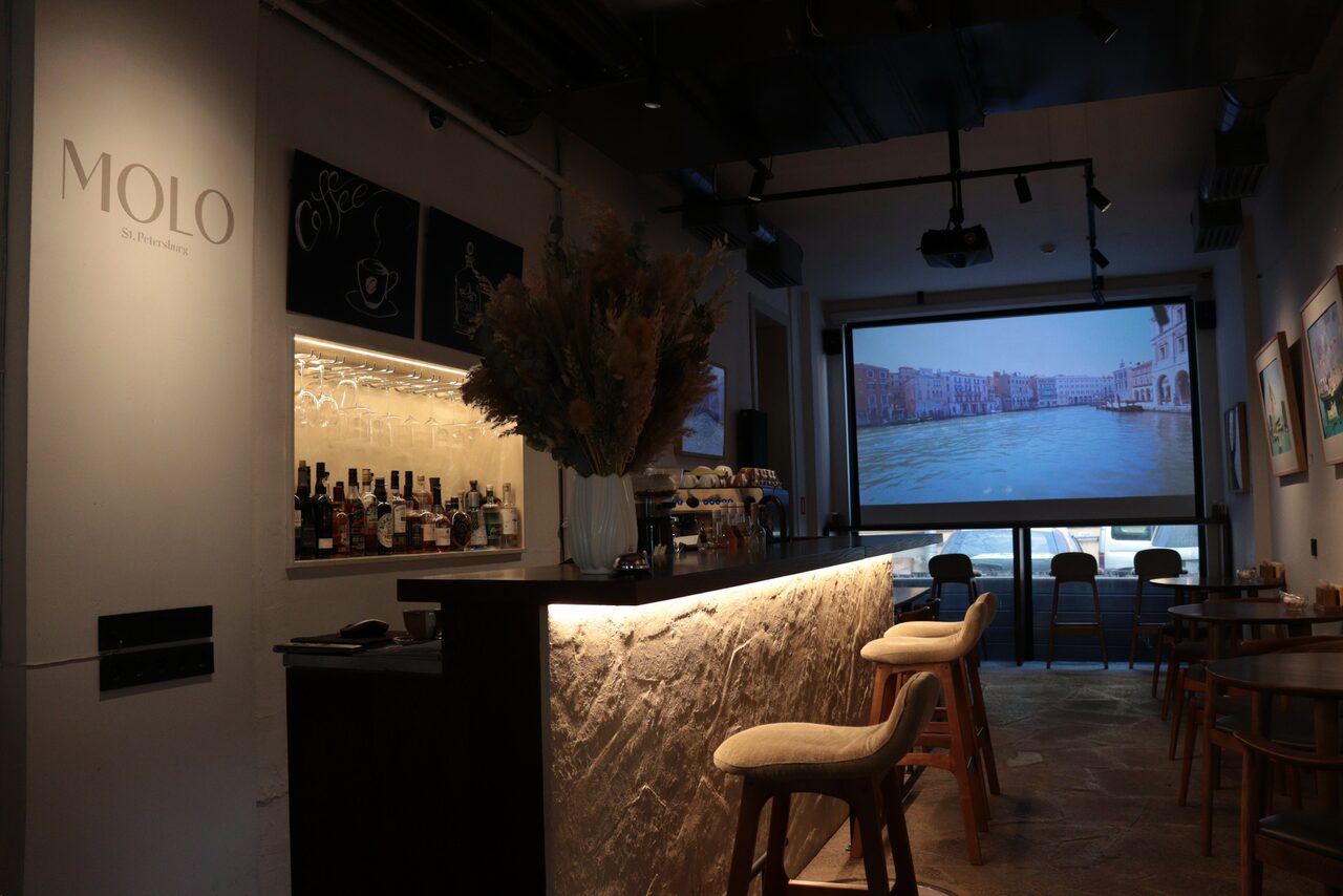Арендовать Гастробар в бутик-отеле MOLO(Моло) 4. Барная зона с центральной стойкой, облицованной светлым камнем, за которой расположены стулья на высоких ножках. За стойкой полки с напитками. На стене проектор с изображением канала. Теплое освещение, потолочные трековые светильники.