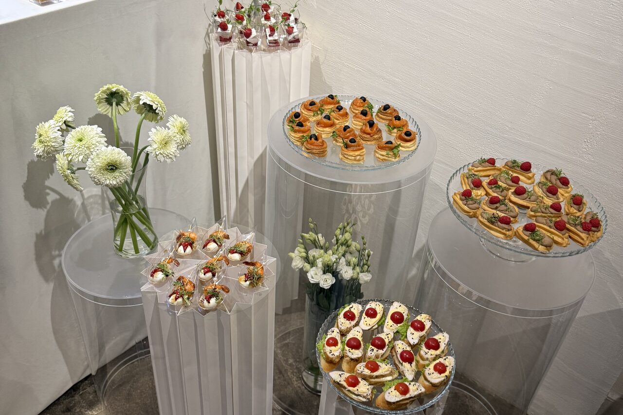 Арендовать Гастробар в бутик-отеле MOLO(Моло) 4. Пространство для мероприятий с белыми стенами, прозрачные цилиндрические столы разной высоты. На столах размещены блюда с канапе и десертами, декорированы цветами в вазах. Общее освещение мягкое, равномерное.