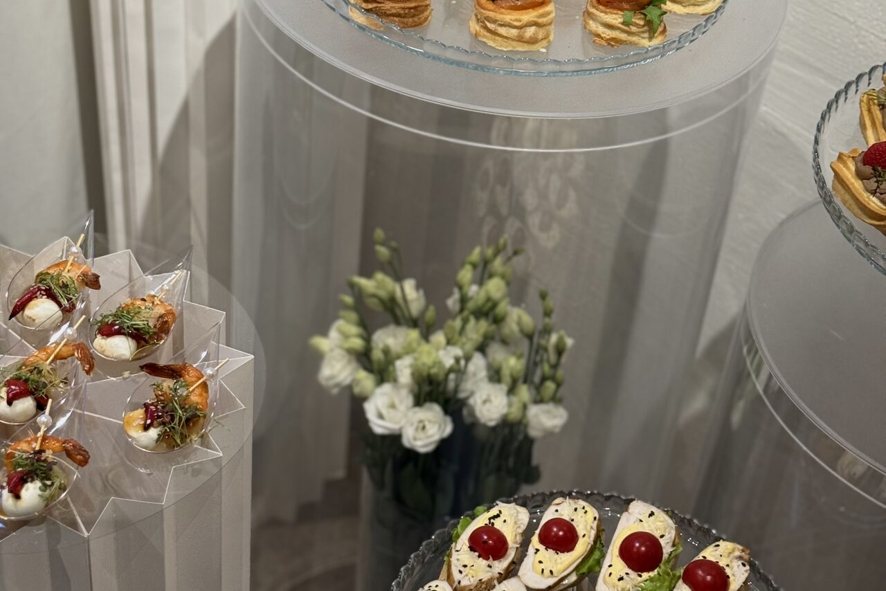 Арендовать Гастробар в бутик-отеле MOLO(Моло) 4. Прозрачные круглые столы с закусками на стеклянных тарелках, закуски в белых и прозрачных контейнерах, в центре ваза с белыми цветами, нейтральная цветовая палитра, столешницы без скатертей.
