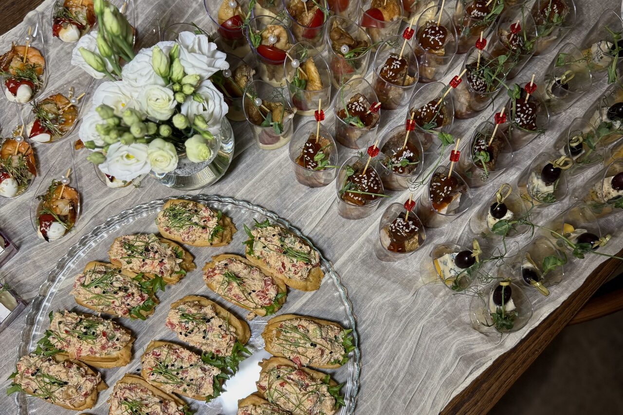 Арендовать Гастробар в бутик-отеле MOLO(Моло) 4. Стол с закусками: бутерброды с зеленью на большом блюде, порционные бокалы с соусом и семенами, фуршетные канапе с оливками. Белый букет цветов. Столовая скатерть нейтрального цвета.