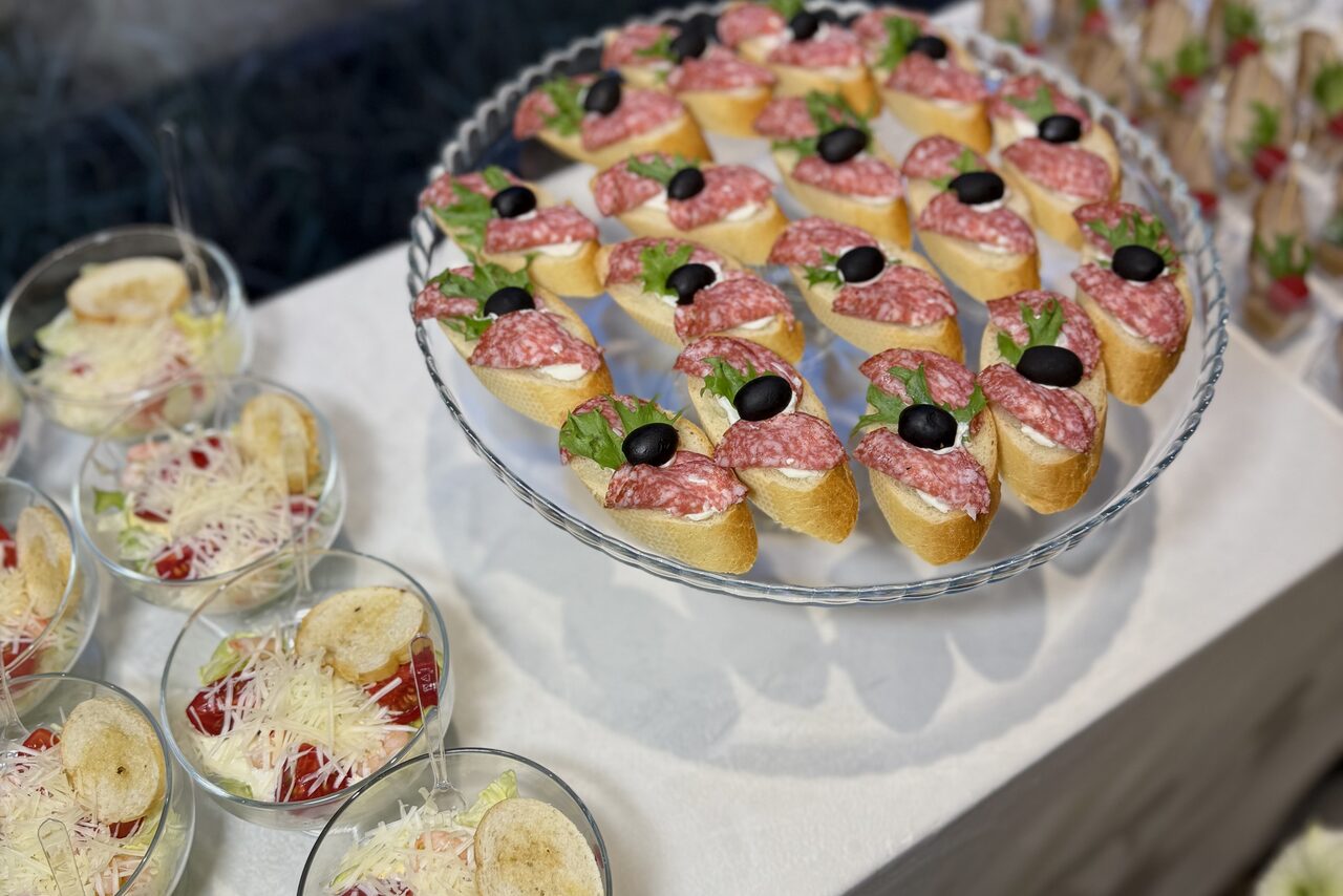 Арендовать Гастробар в бутик-отеле MOLO(Моло) 4. Разнообразные закуски на светлой скатерти: на стеклянной тарелке бутерброды с салями, зеленью и оливками, рядом прозрачные бокалы с салатом, посыпанным сыром.