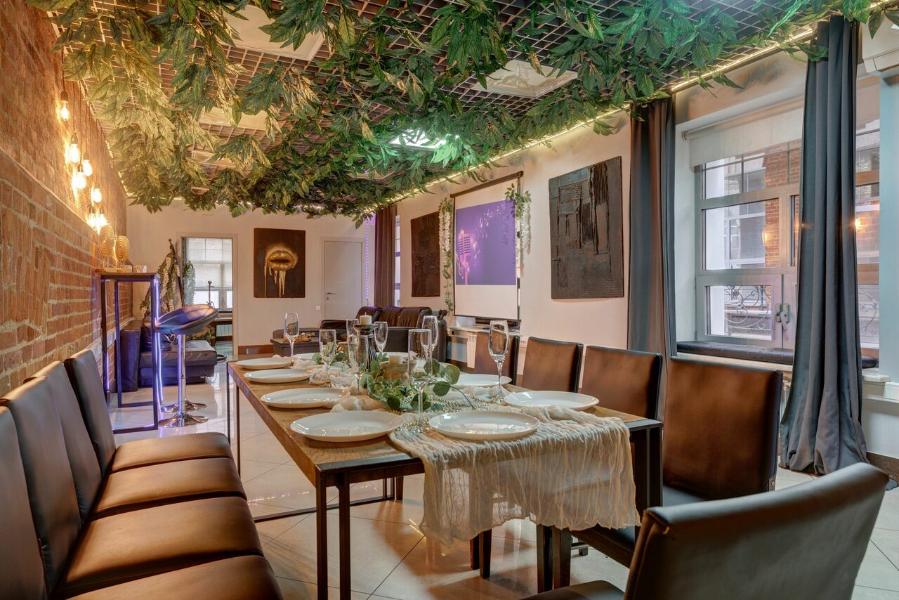 Арендовать Лофт Семеновский. Пространство с кирпичной стеной и подвесным декором из зелени. Длинный обеденный стол с сервировкой на восемь человек, кожаные стулья. Окна с темными шторами. Потолочные светильники, светлые плиточные полы, проектор и экран.