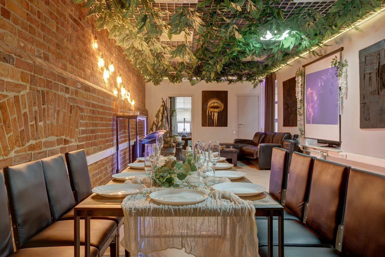 Арендовать Лофт Семеновский. Длинный стол с сервировкой и черными стульями, кирпичная стена слева, потолок украшен зелеными растениями, неоновая подсветка, на стене картины и проектор, сзади диваны, оформлено в теплых тонах, есть декоративные растения.