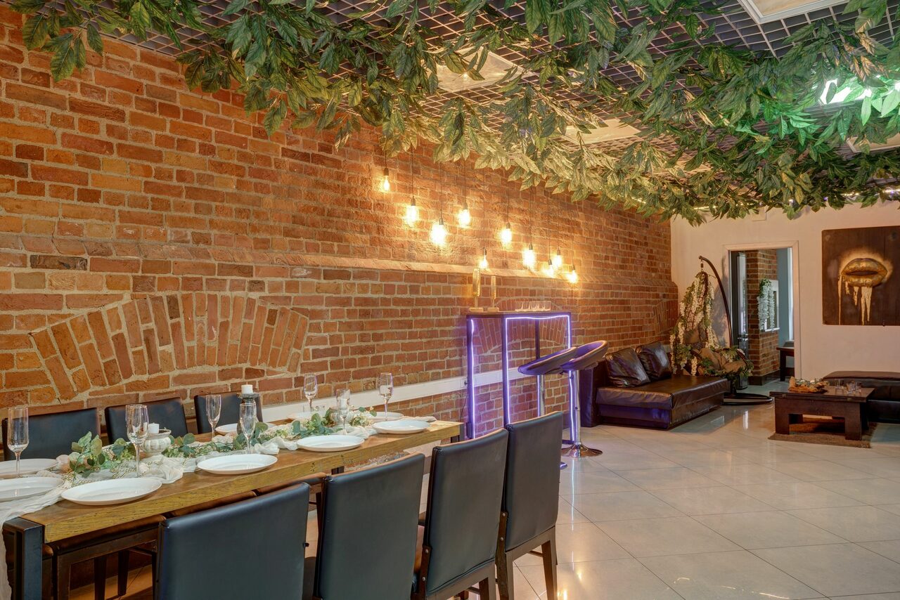 Арендовать Лофт Семеновский. Ресторанное пространство с кирпичными стенами и подвесной декорацией из зелени. Длинный стол, сервированный тарелками и бокалами, с черными стульями. Мягкий диван в углу. Настенные светильники, картина. Пол выложен плиткой.