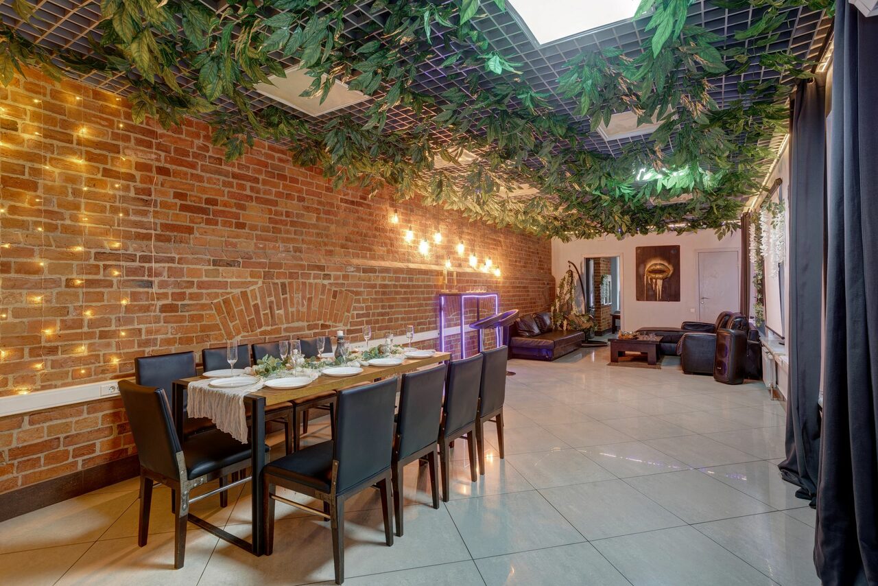 Арендовать Лофт Семеновский. Просторное помещение с кирпичной стеной, вдоль которой стоит накрытый стол и десять стульев. Потолок украшен зеленью, пол выложен светлой плиткой. Освещение представлено гирляндами и лампами. В дальней части виден кожаный диван.