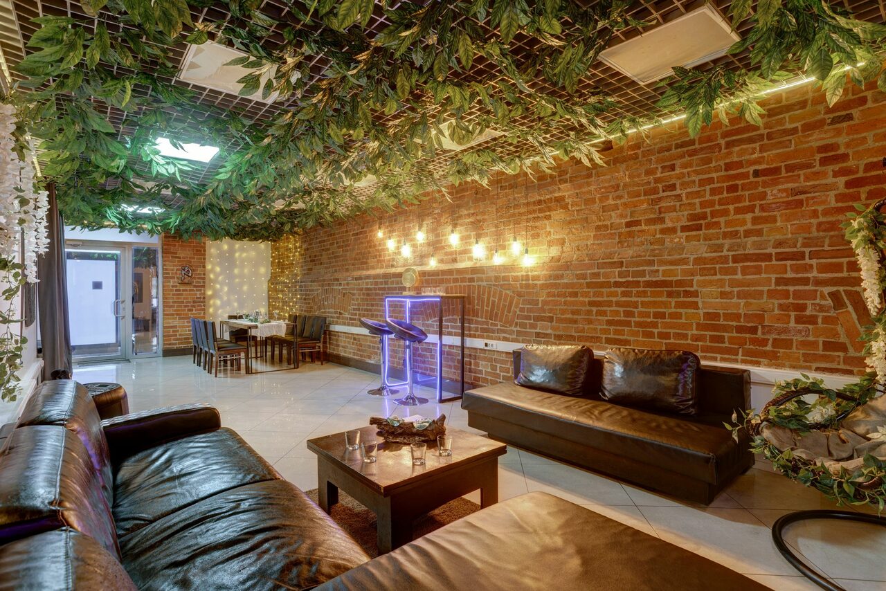 Арендовать Лофт Семеновский. Лофт-пространство с кирпичными стенами. Диваны и столы из темного дерева. Потолок с зелеными декоративными растениями. Подвесные светильники на стене. Плиточный пол светлого цвета. Стойка с подсветкой. Планировка с зонами для отдыха и приема пищи.