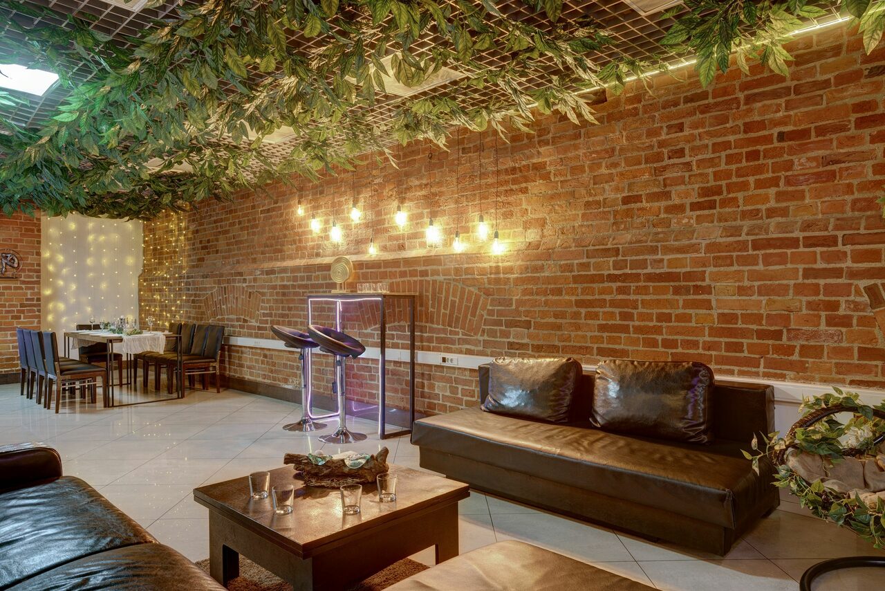 Арендовать Лофт Семеновский. Пространство с кирпичными стенами, декорированными гирляндами. На потолке искусственная зелень. Черные кожаные диваны, деревянный стол. Светильники с плафонами. Зона с микрофонами и стойкой для караоке. На полу светлая плитка. Стол с черными стульями.