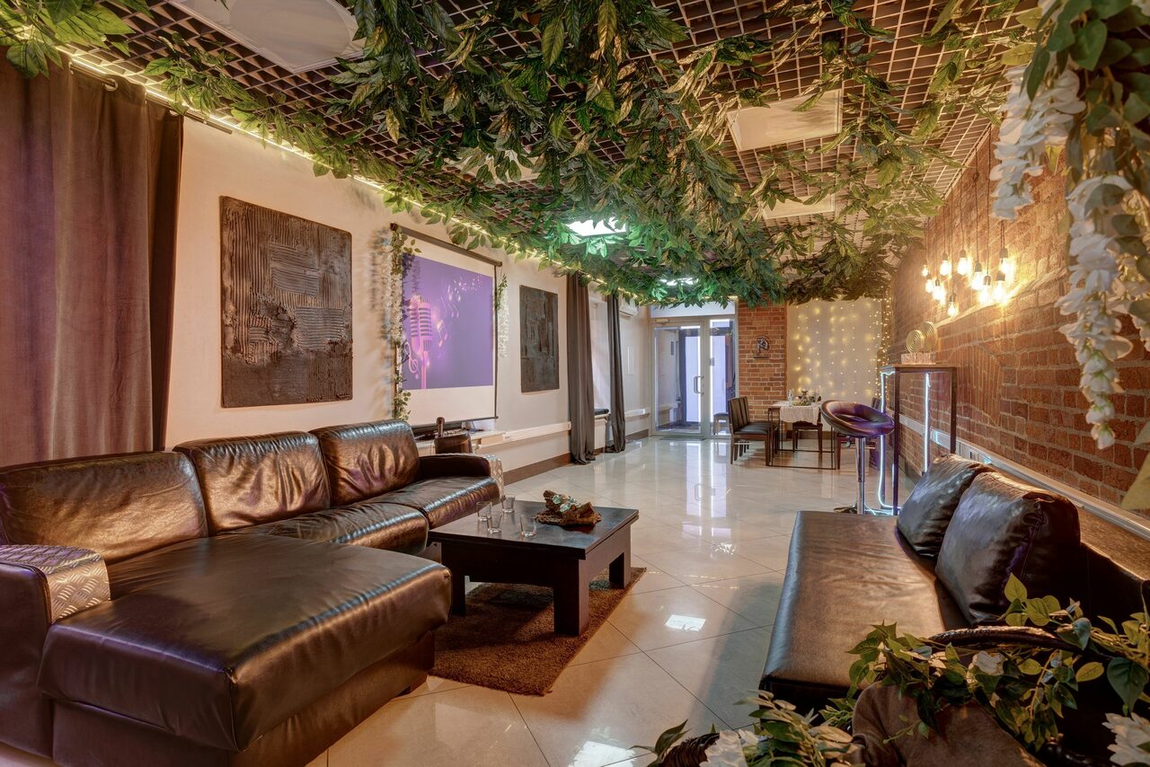 Арендовать Лофт Семеновский. Просторная комната с кожаными диванами, темным журнальным столиком и барной зоной. Плиточный пол, кирпичная стена, потолок с декоративной растительностью. Теплое освещение, картины на стенах, фиолетовый экран сбоку. Комнатные растения добавляют уюта.