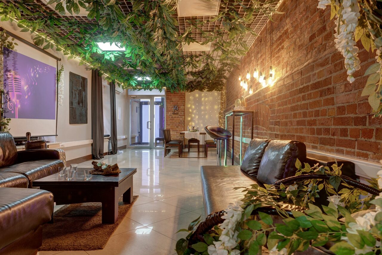 Арендовать Лофт Семеновский. Просторное помещение с ярким освещением, стены с кирпичной кладкой, зеленая декоративная растительность на потолке. Темная кожаная мебель, небольшой стол, барная стойка. Светлая плитка на полу, на стене экран с изображением.