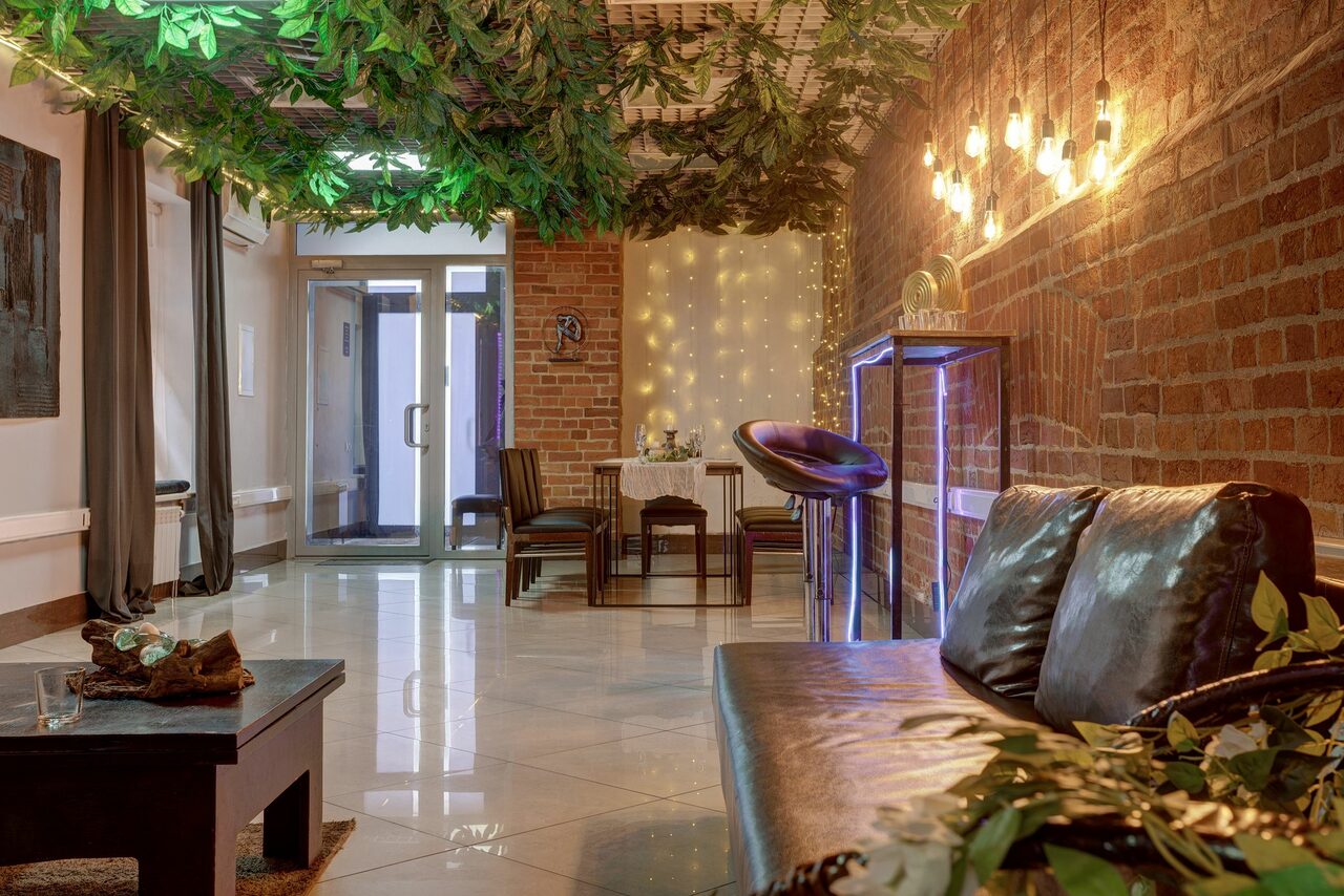 Арендовать Лофт Семеновский. Лофт-пространство с кирпичными стенами и глянцевым полом. Потолок украшен искусственными листьями. Мягкий диван, стол с креслами, аквариум. Настенные светильники и гирлянда создают атмосферное освещение. Входная дверь из стекла.