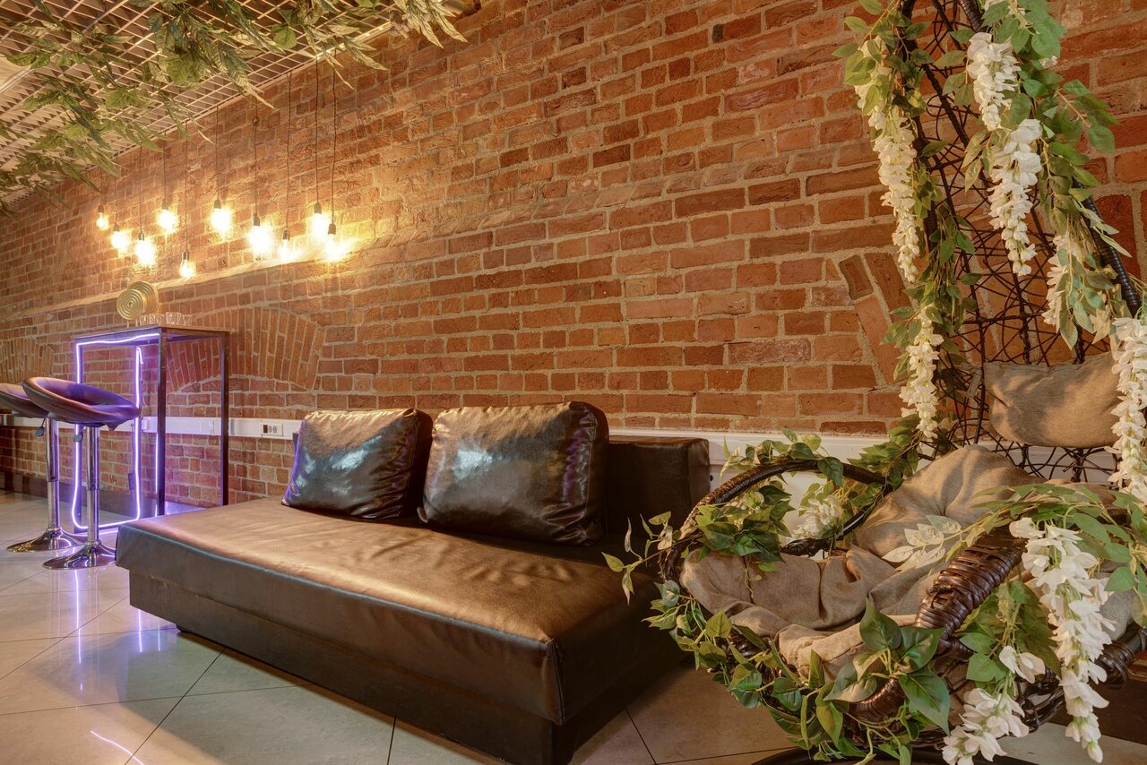 Арендовать Лофт Семеновский. Лофт-пространство с кирпичными стенами и плиточным полом. Коричневый кожаный диван, плетеное кресло с белыми цветами и растениями. Подвесные светильники, барный стул, неоновая стойка. На потолке декоративные растения.