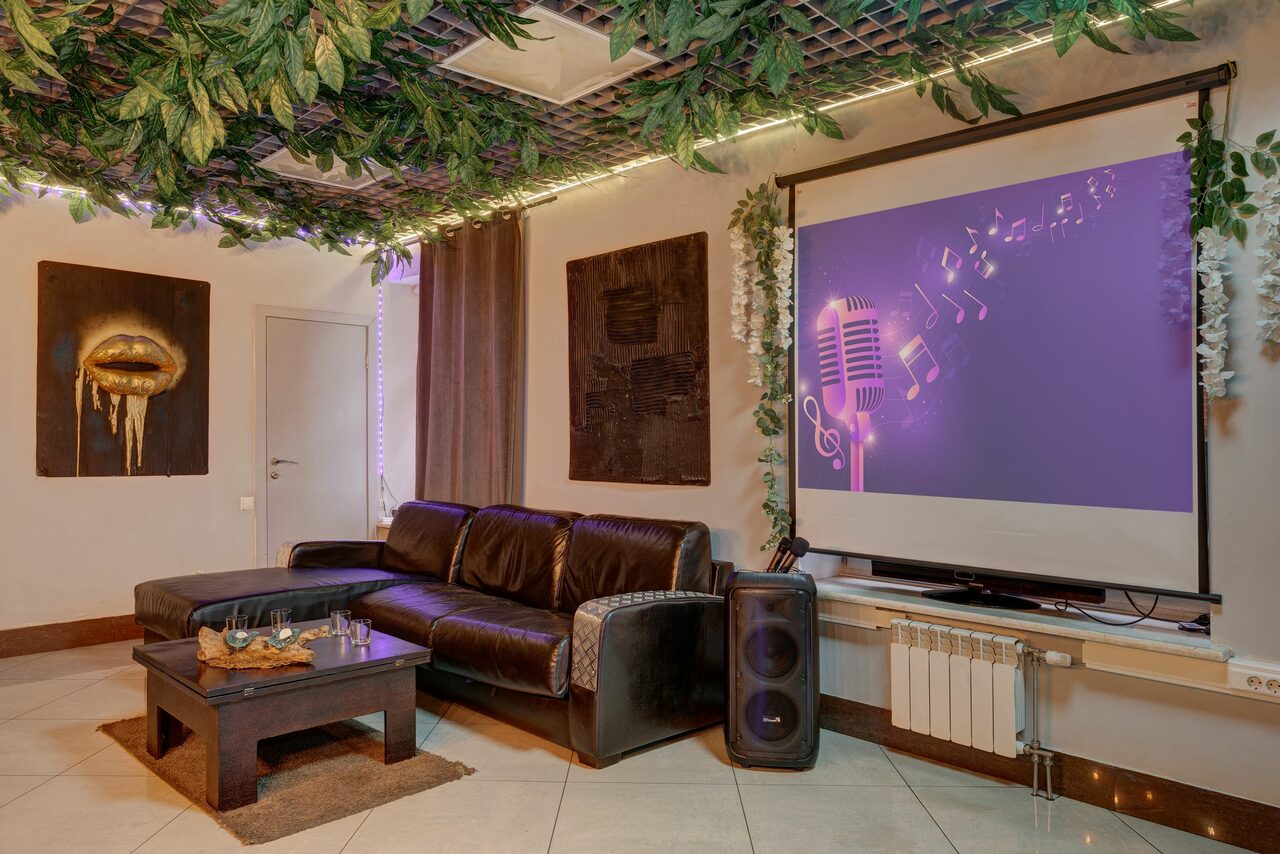 Арендовать Лофт Семеновский. Пространство с диваном, кофейным столиком и проектором на стене. Потолок украшен искусственными растениями и подсветкой. На стенах картины, справа колонка и батарея. Пол плиточный, на столе поднос с питьем. Цвета: коричневый, белый, зелёный.