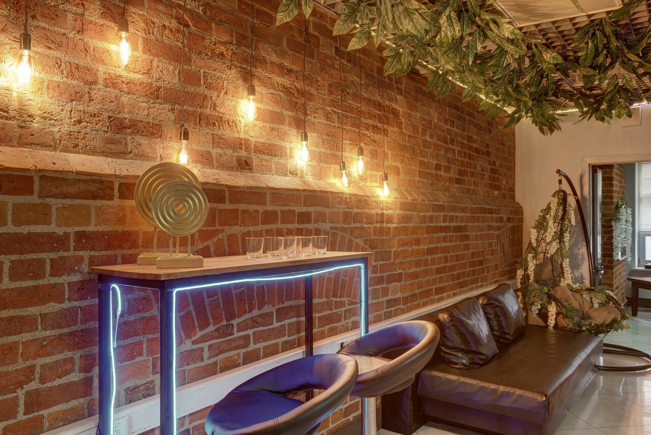 Арендовать Лофт Семеновский. Лофт-пространство с кирпичными стенами, подвесными лампами, барной стойкой с подсветкой, двумя барными стульями, черным диваном, декоративной зеленью на потолке. Уютный интерьер для мероприятий.