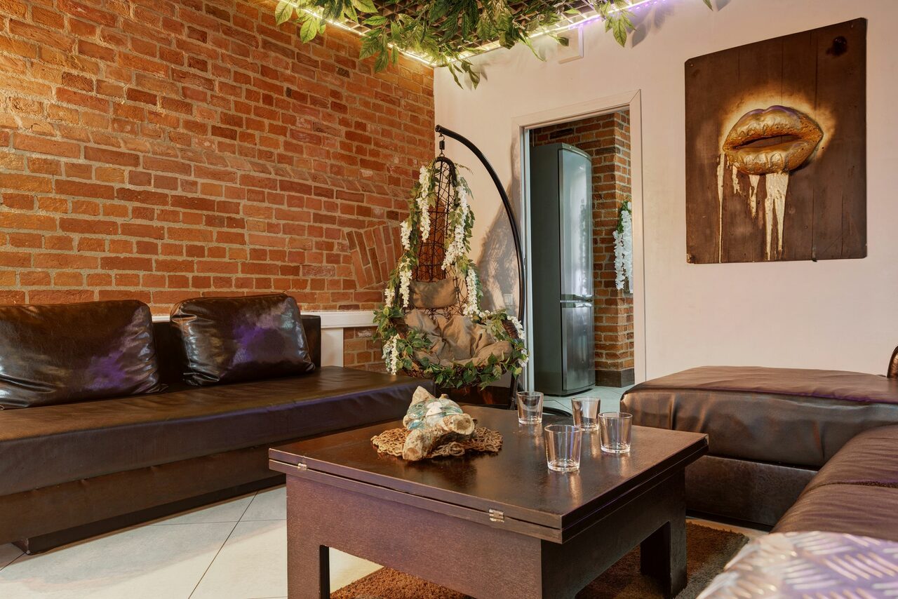 Арендовать Лофт Семеновский. Гостиная с коричневыми кожаными диванами и деревянным столом, на котором стоят стаканы и декор. Стена из красного кирпича, белая стена с картиной губ. Подвесной декор с зеленью. Видна дверь в другое помещение.
