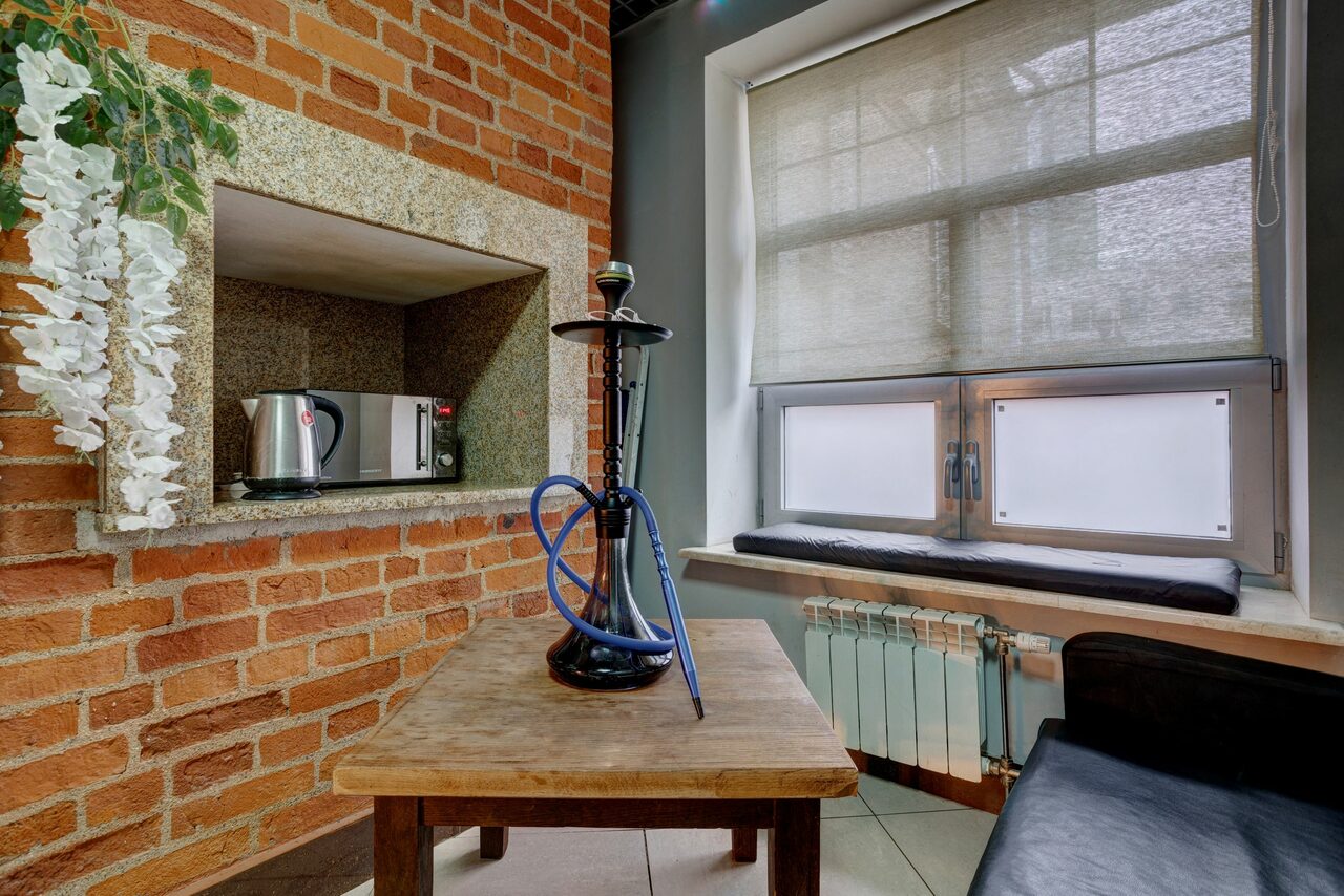 Арендовать Лофт Семеновский. Помещение с кирпичной стеной, нишей с микроволновкой, деревянным столом и кальяном. Большое окно с жалюзи, подоконник с мягкой обивкой. Черный стул, радиатор отопления. Светлые плиточные полы, искусственные растения на стене.