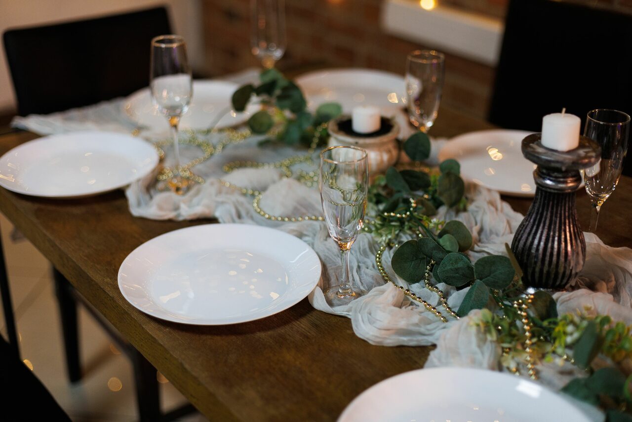 Арендовать Лофт Семеновский. Деревянный обеденный стол с белыми тарелками, бокалами и декором из зелени и тканей. На столе расположены декоративные свечи. На заднем плане кирпичная стена. Стулья черные. Освещение мягкое.