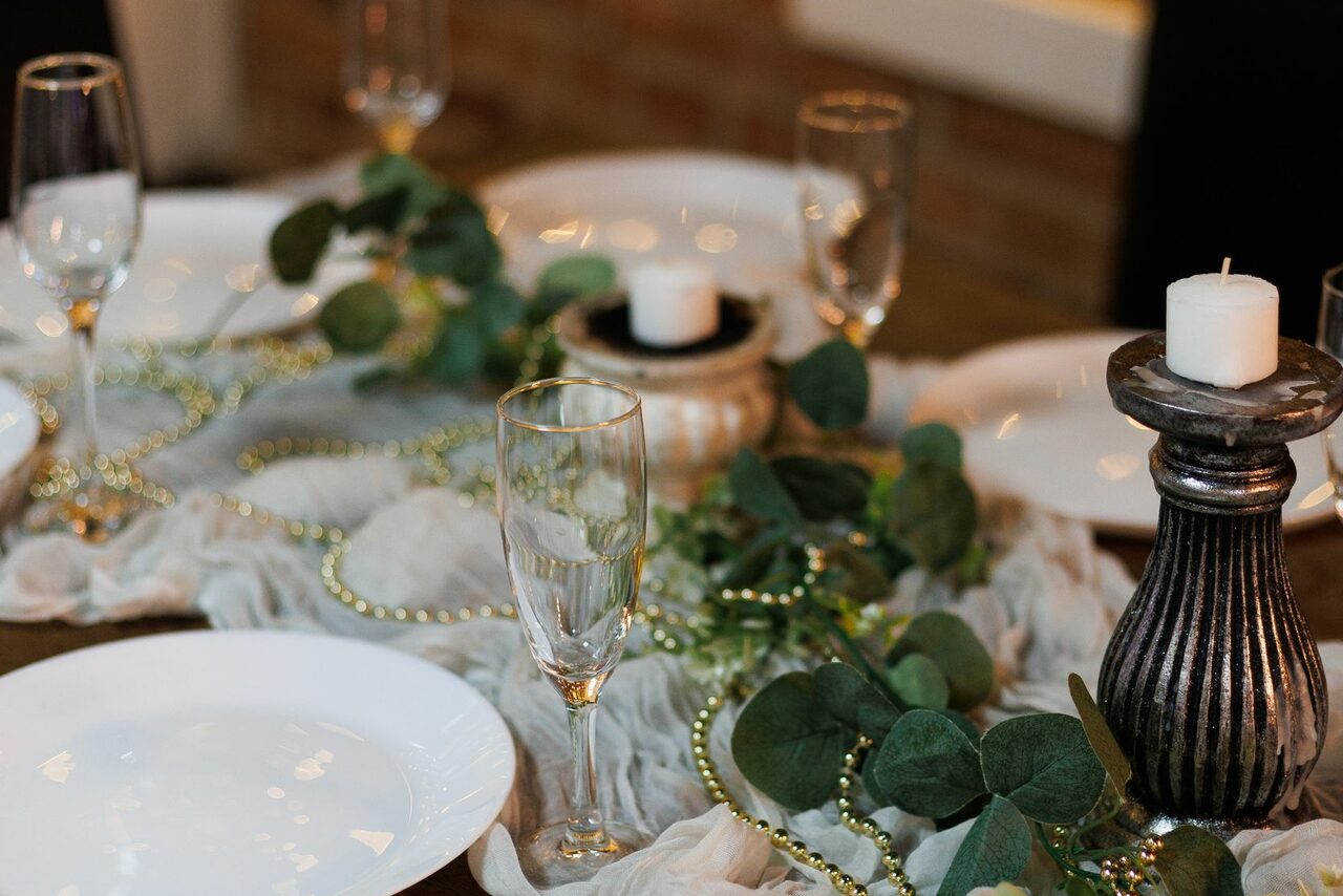 Арендовать Лофт Семеновский. Стол с белыми тарелками, бокалами и свечами в подсвечниках. По центру расположена композиция из зелёных веток и золотистых бус. Фон кирпичный. Освещение тёплое.