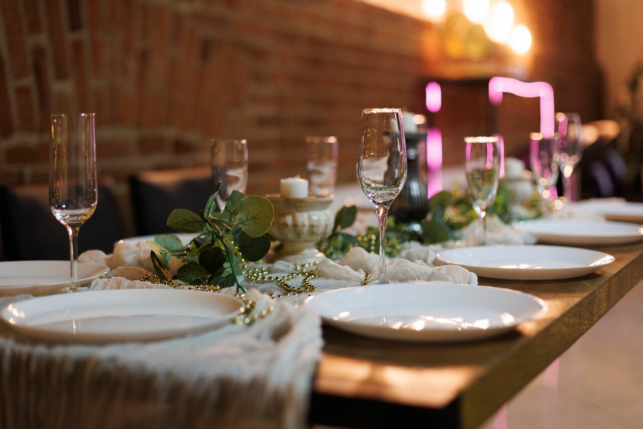 Арендовать Лофт Семеновский. Длинный деревянный стол с сервировкой на шесть мест: тарелки, бокалы, столовые приборы. Декор из зелени и свечей. Фоновая стена из кирпича, мягкое освещение, светодиоды.