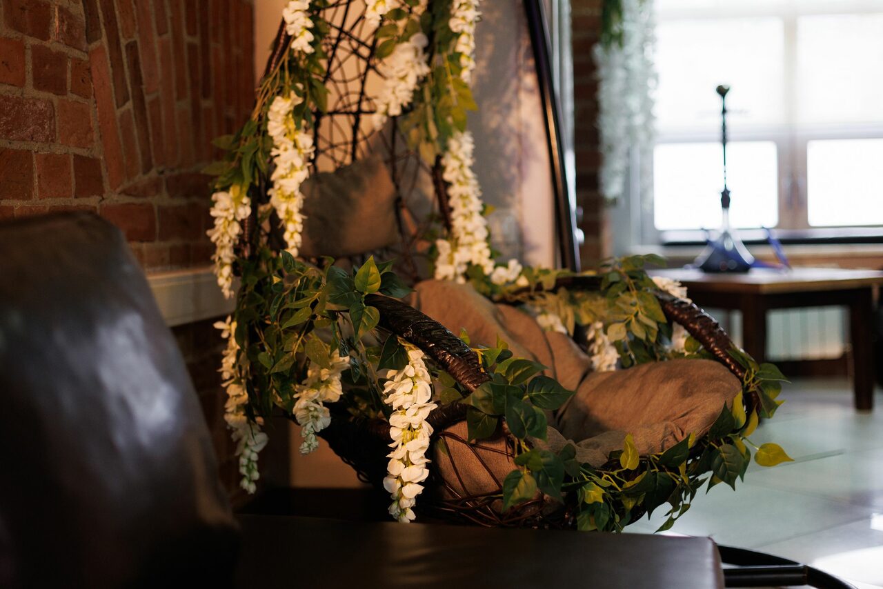 Арендовать Лофт Семеновский. Лофт-пространство с кирпичной стеной, подвесное кресло с растительными декорациями и подушками, кожаное кресло, стол с лампой у окна. Естественное освещение, светло-серые и коричневые оттенки, деревянные и текстильные материалы.