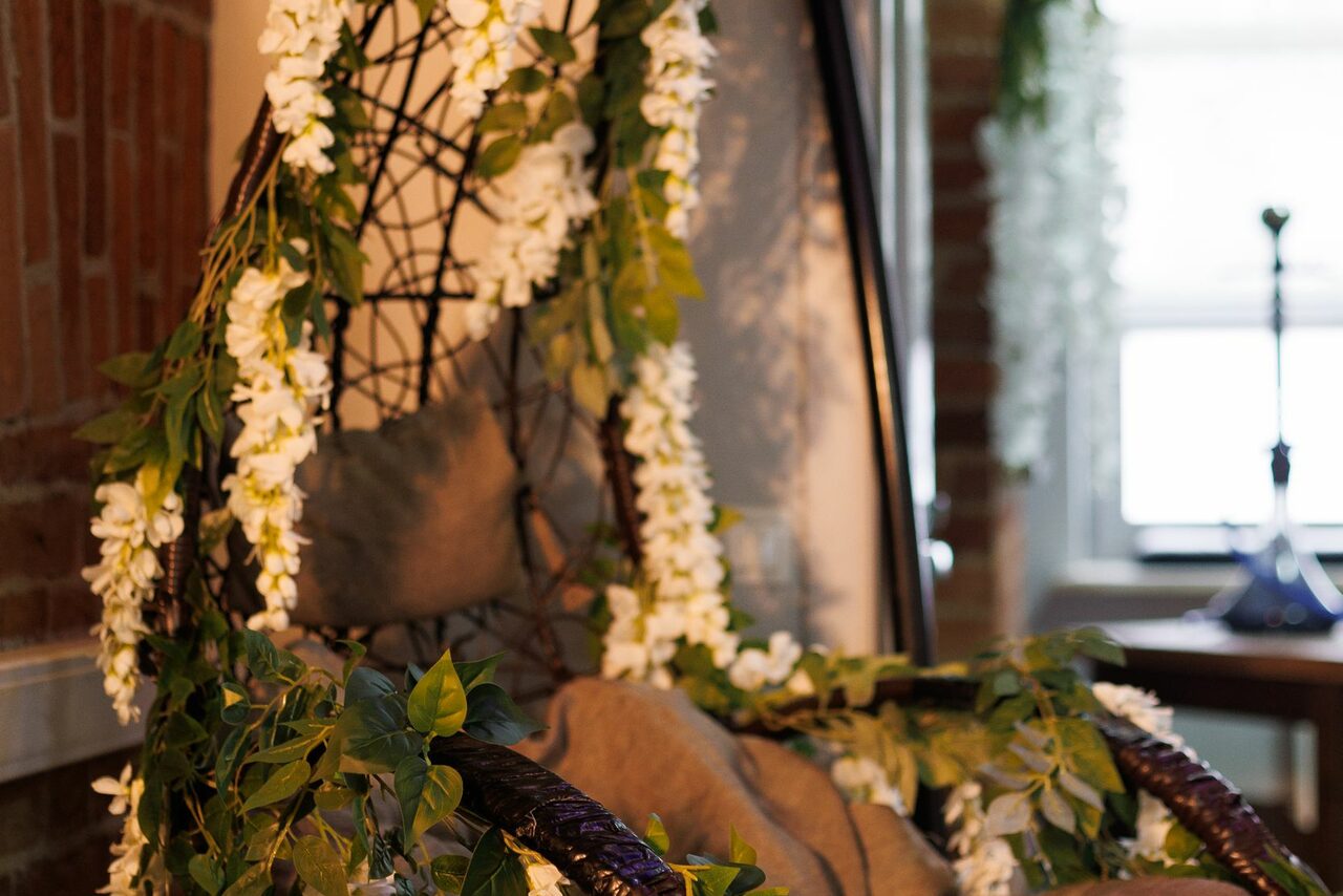 Арендовать Лофт Семеновский. Подвесное плетёное кресло на фоне кирпичной стены, украшенное белыми цветами и зелёной листвой, с подушкой и покрывалом. Естественное освещение через окно. Интерьер в стиле лофт.