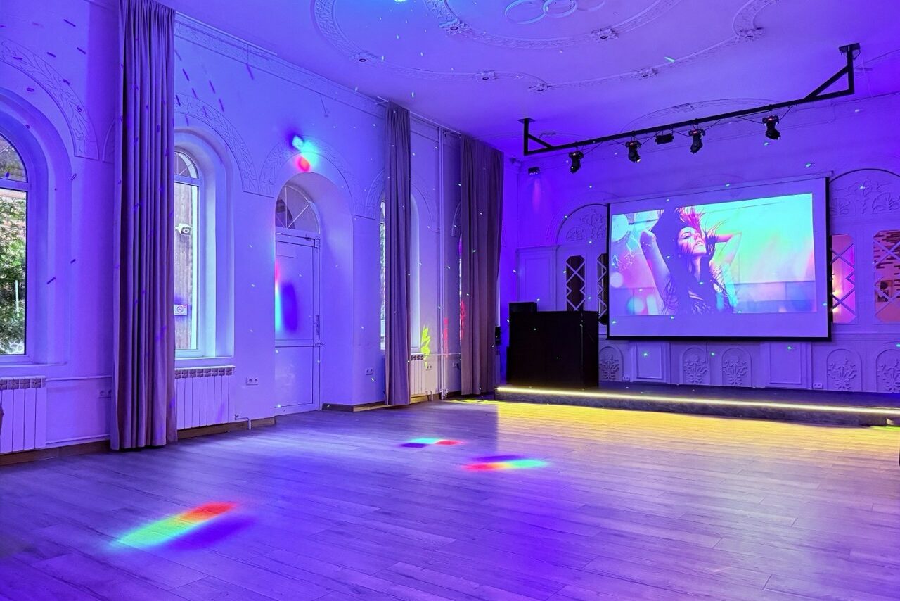 Арендовать Лофт Софт. Просторное помещение с высокими потолками, светлой деревянной плиткой на полу. Имеет большие арочные окна с занавесками. На стене висит экран с изображением, рядом черная аудиосистема. Освещение разноцветное, создающее яркие лучи на полу.