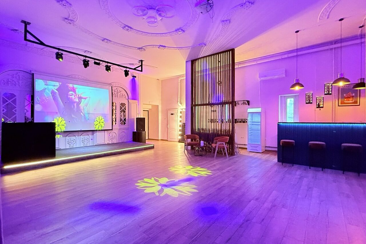 Арендовать Лофт Софт. Просторное помещение с деревянным светлым полом, сценой, подсветкой фиолетового и желтого цветов. Справа — барная стойка с высокими стульями. На стене — большой экран с проектором. Потолок украшен лепниной, подвесные лампы над баром.