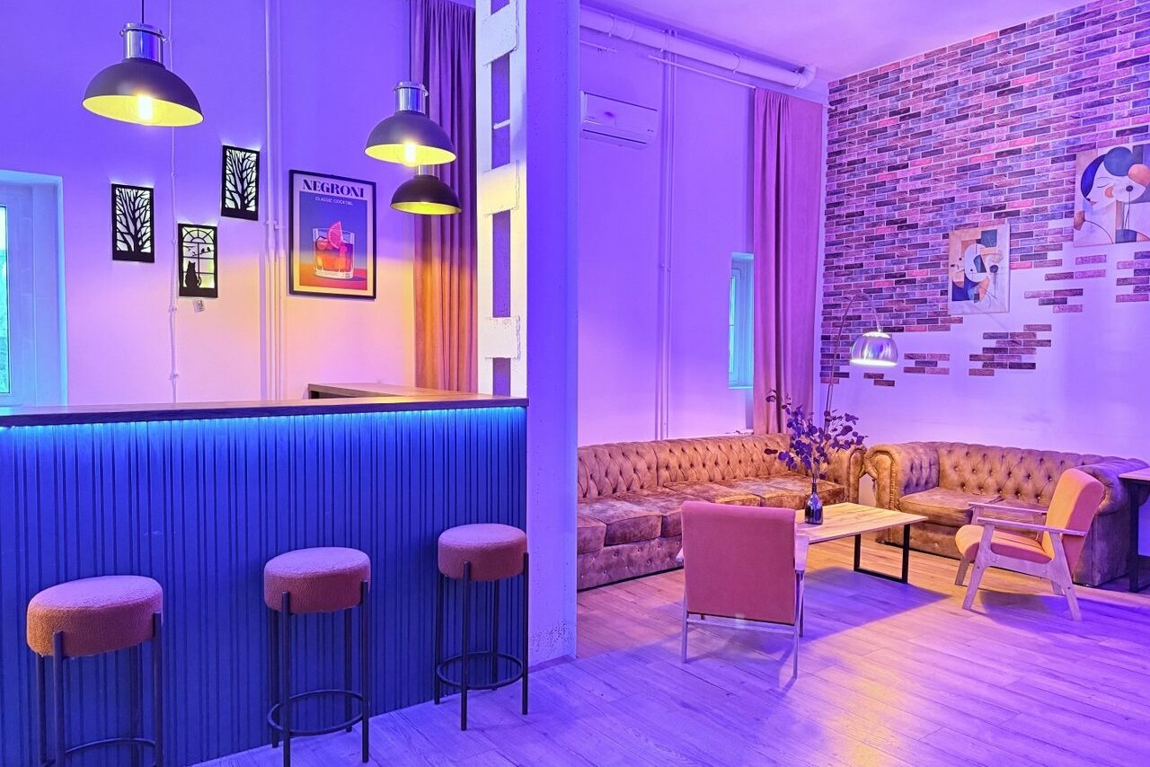 Арендовать Лофт Софт. Лофт-пространство с барной стойкой и тремя стульями, мягкий диван, кресло и стол. Яркое освещение, разноцветные неоновые акценты, кирпичная стена с картинами, светлый деревянный пол, фиолетовые шторы. Настенные светильники и подсветка стойки.