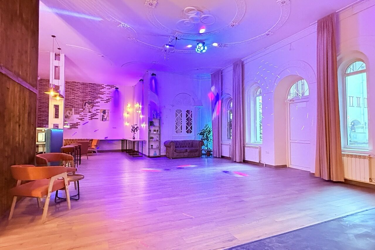 Арендовать Лофт Софт. Просторный зал с деревянным полом и высокими потолками. Стены белые с декоративными элементами. Освещение разноцветное, мягкое. По периметру стоят стулья и диваны. Есть арочные окна с занавесками. На стенах декор из кирпича и зеркала.