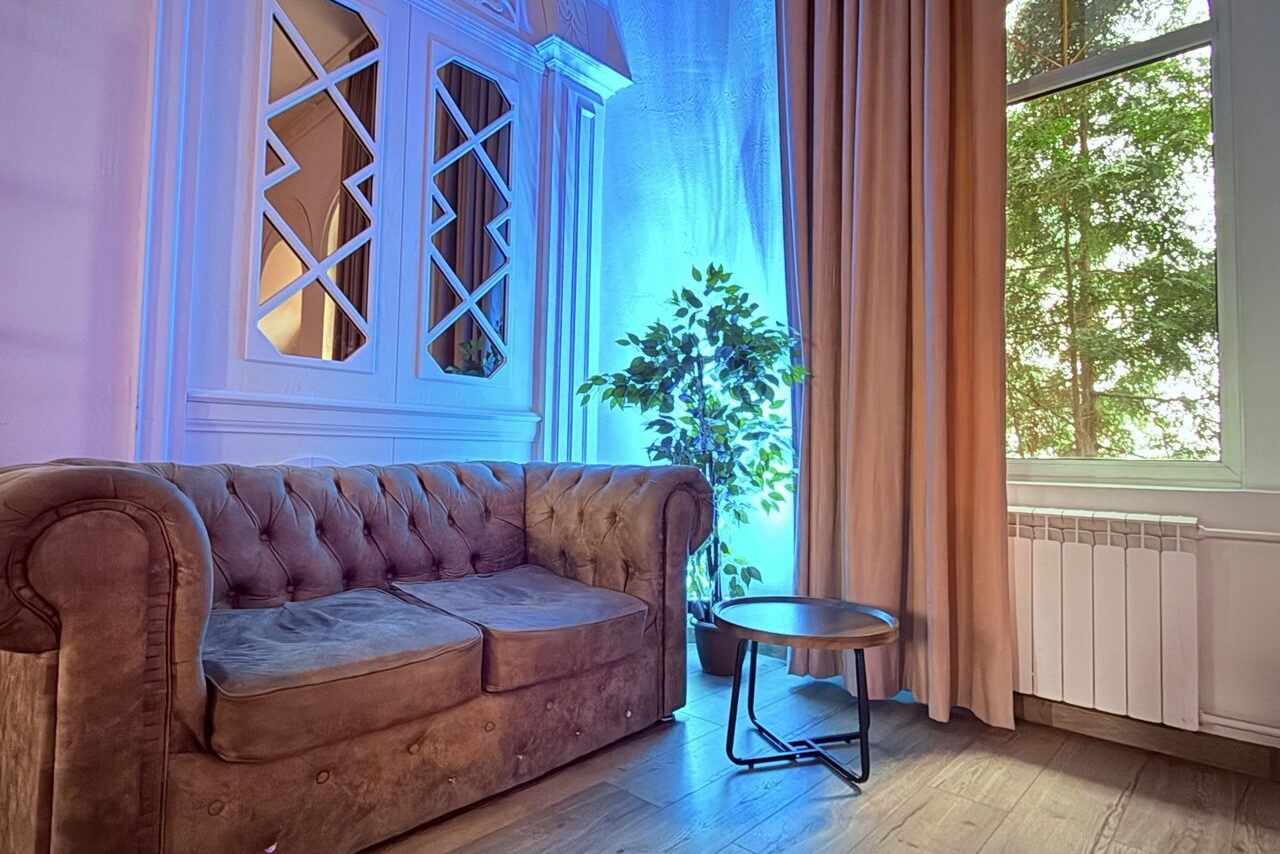 Арендовать Лофт Софт. Гостиная с коричневым диваном, круглым кофейным столиком и растением в горшке у окна. Крупные шторы, радиатор, пол паркетный. Нейтральные цвета, стены украшают зеркала с геометрическим узором. Синяя декоративная подсветка.