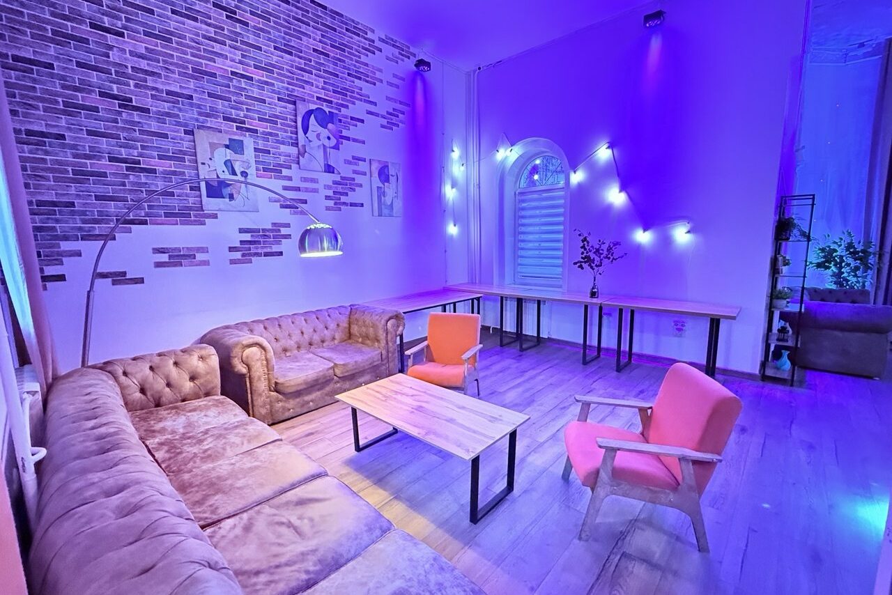 Арендовать Лофт Софт. Просторная гостиная с кирпичной стеной, фиолетовым освещением, двумя мягкими диванами и двумя креслами, деревянный стол. Белые стены и пол, большие окна, декоративные растения и картины. Светодиодная гирлянда вдоль стены.