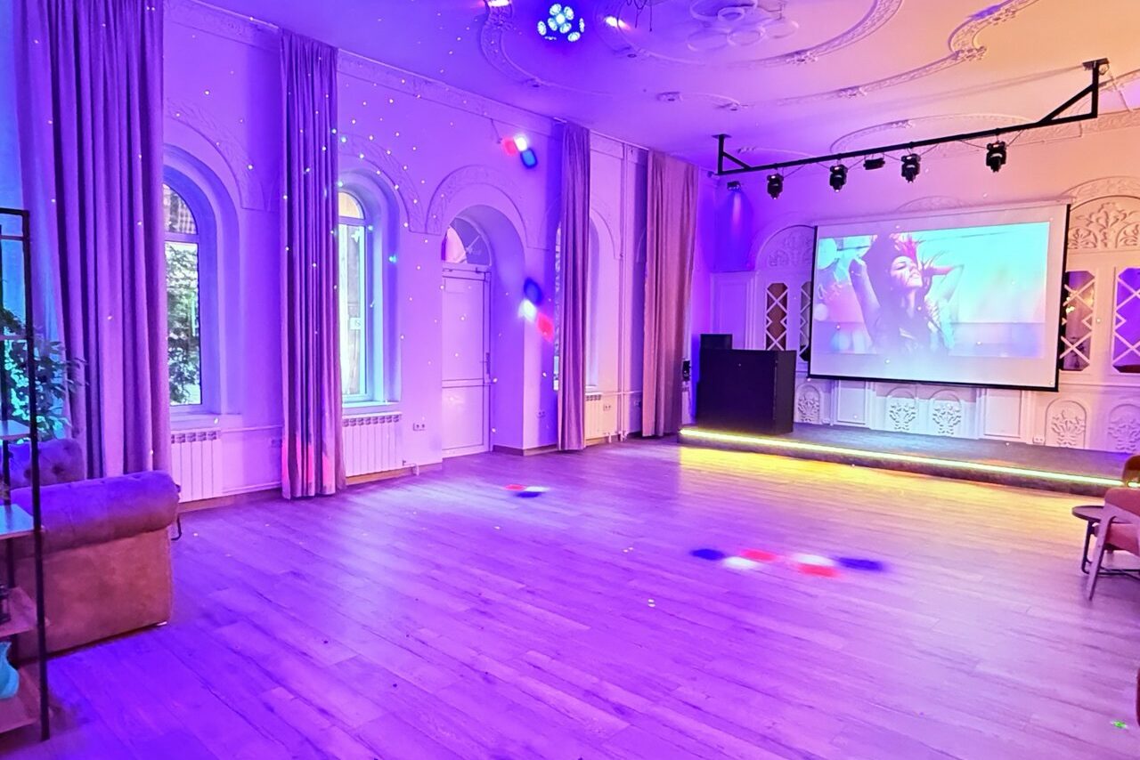 Арендовать Лофт Софт. Просторное помещение с деревянным полом, арочными окнами и шторами. На стене проектор с изображением, рядом колонка. Потолочные светильники и разноцветные световые эффекты. В углу мягкий диван. Белые стены с декоративными элементами.
