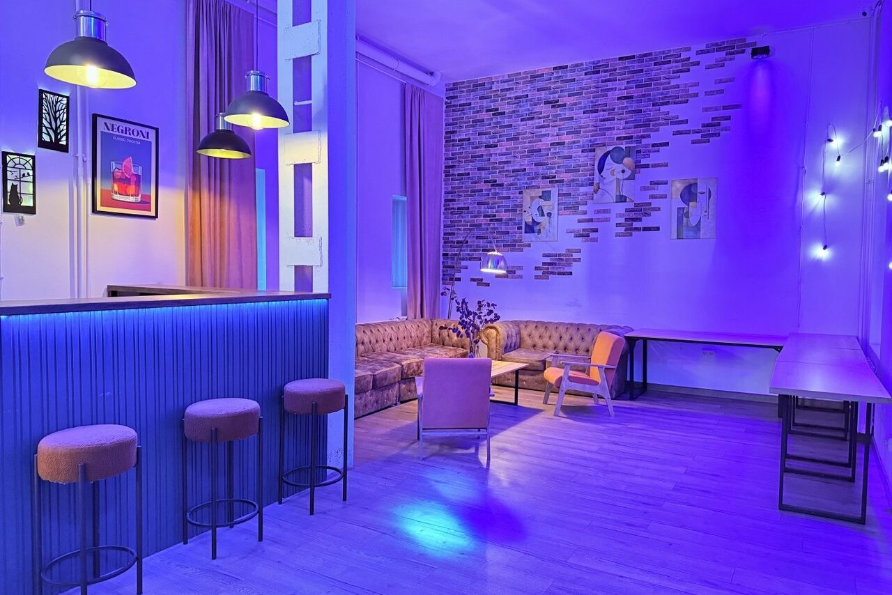 Арендовать Лофт Софт. Loft-пространство с барной стойкой и тремя стульями, мягкий уголок с диванами и креслом, настенные полки, декоративная кирпичная стена, яркая неоновая подсветка, подвесные светильники, длинный рабочий стол.