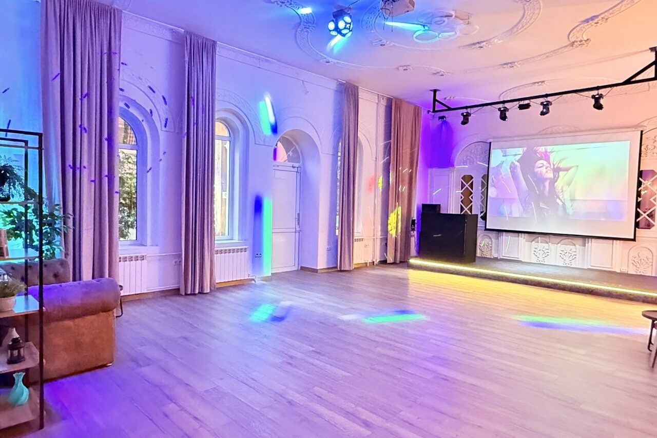 Арендовать Лофт Софт. Просторное помещение с деревянным полом и высокими окнами с занавесками. На стене установлен большой экран с изображением, присутствует проигрывательное оборудование. Потолочное освещение разноцветное, на стенах декоративные элементы.