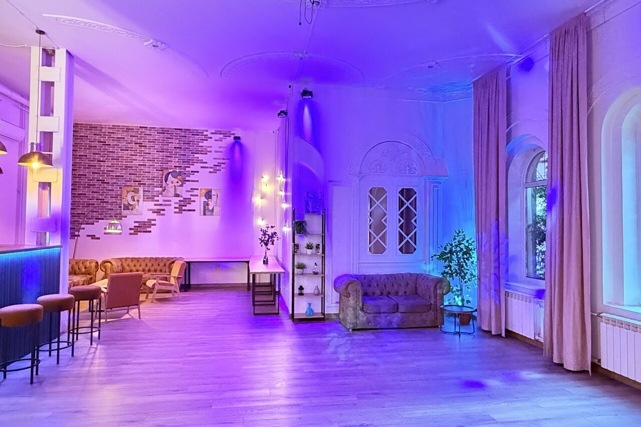 Арендовать Лофт Софт. Просторное помещение с барной стойкой и высокими стульями справа, зона отдыха с мягкими диванами и столиком слева. На стенах кирпичные и светлые отделки, неоновая подсветка. Окна с длинными шторами. Паркетный пол.